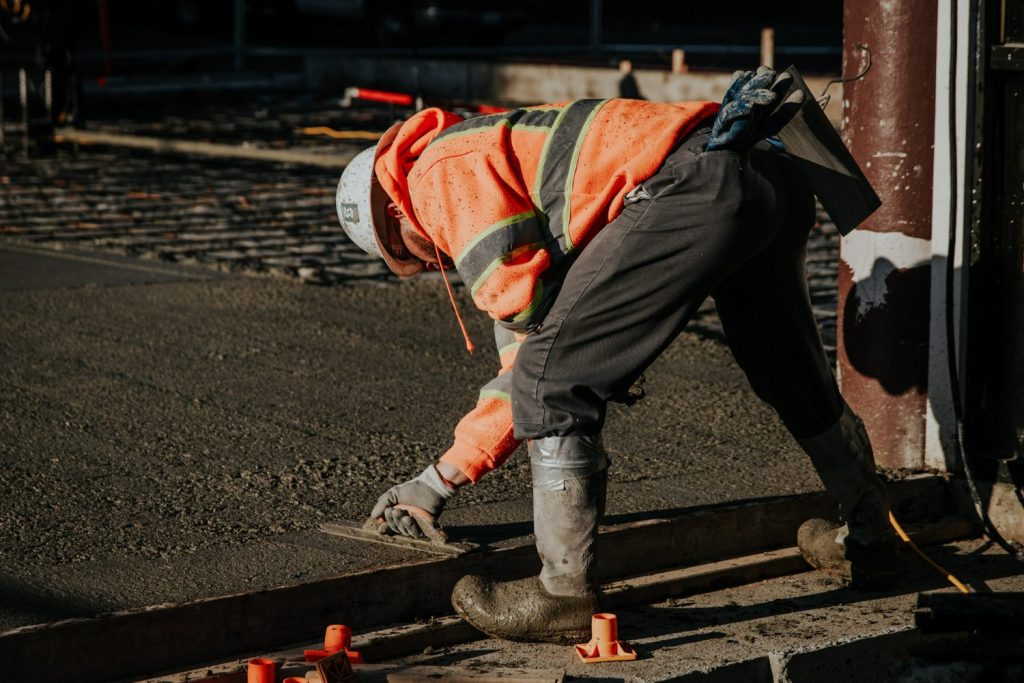 Trabajador de la construcción con chaleco reflectante naranja y casco alisando hormigón con una llana